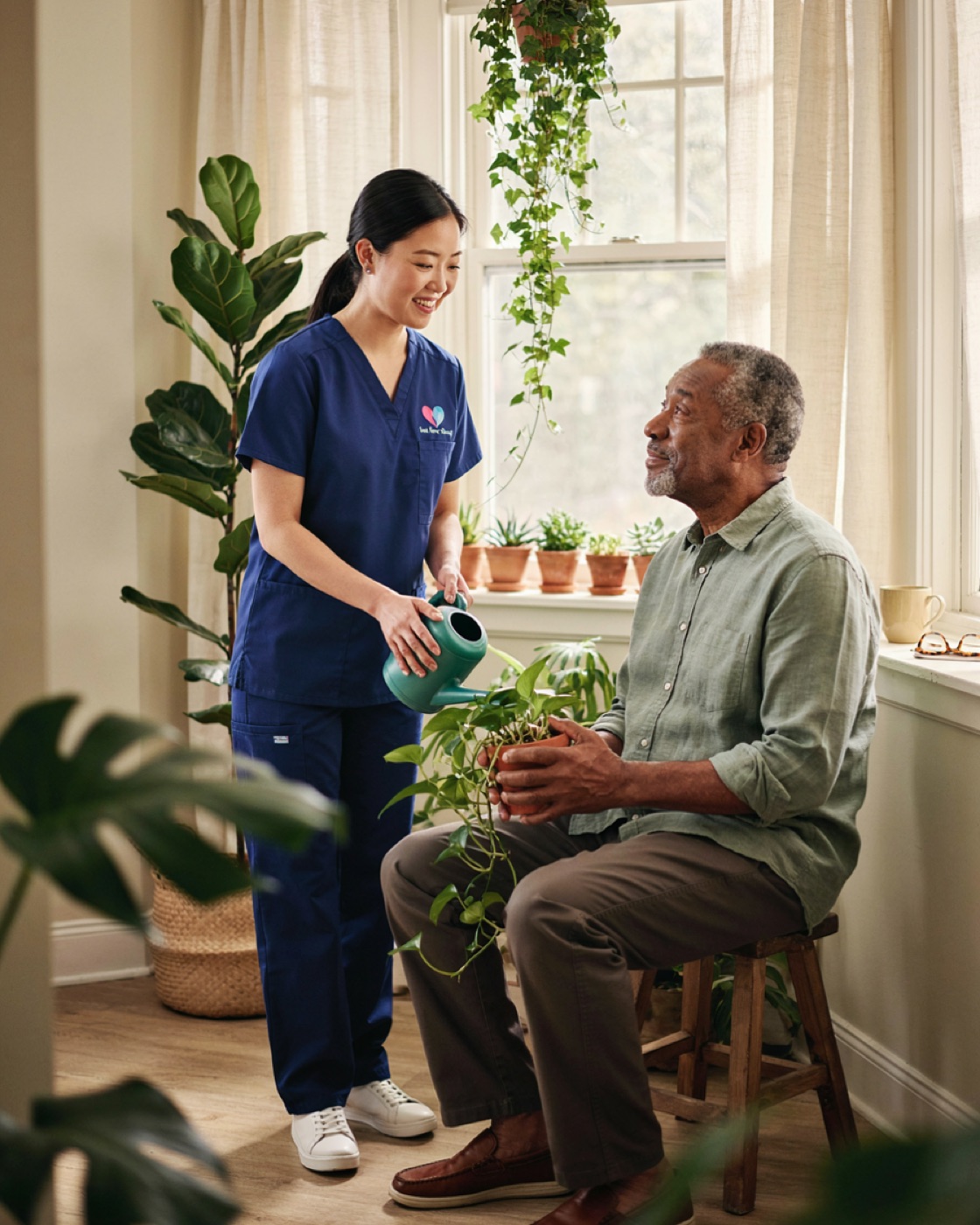 A caregiver in scrubs helping an older man water his houseplants by a sunny window